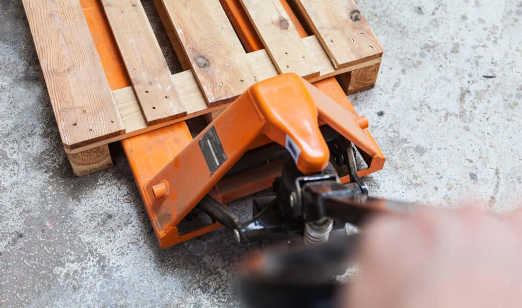Vue rapprochée de l'avant d'un transpalette manuel orange soulevant une palette en bois, sur sol en béton brut.