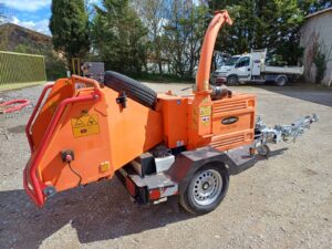 Broyeur de végétaux orange sur remorque, modèle LOCATOP31 TW 1901DMB, avec roue de secours, stationné sur gravier près d'arbres.