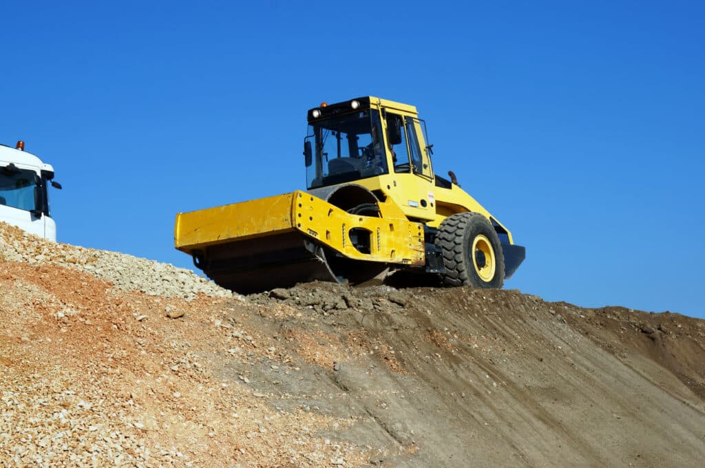 Compacteur vibrant jaune sur une pente de terre et gravats, ciel bleu clair. Cabine de camion blanc visible à gauche.