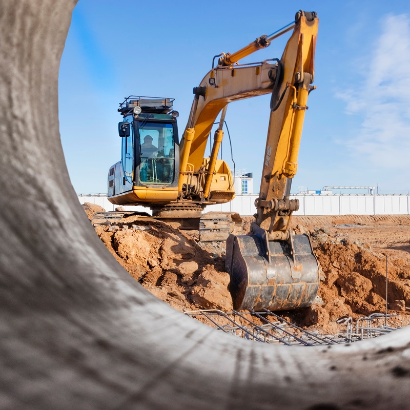 Excavatrice jaune creusant un site de construction avec une armature métallique visible, vue à travers une grande canalisation en béton.