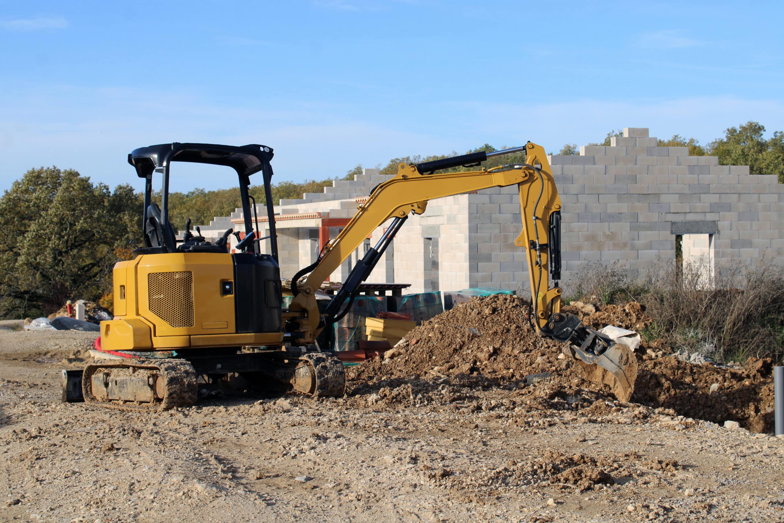 Mini-pelle jaune creusant un tas de terre sur un chantier de construction avec des blocs de parpaings en arrière-plan.