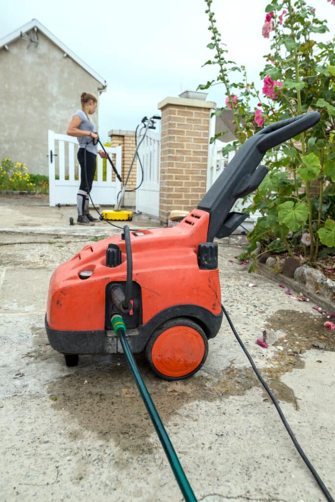 Nettoyeur haute pression orange posé au sol pendant qu'une femme nettoie l'allée en béton.
