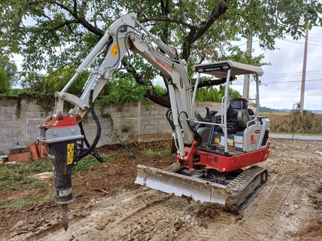 Mini-pelle Takeuchi TB225 avec marteau-piqueur hydraulique Bobcat sur un chantier boueux, devant un mur de parpaings et sous un arbre feuillu.